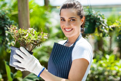 A young woman with dark hair tied back, smiling and wearing a white T-shirt, blue striped apron, and white gardening gloves, holding a potted shrub with small green leaves and pink-tinged edges. She is standing outdoors in a well-maintained garden or greenhouse with vibrant green foliage, including trees, bushes, and flowering plants, indicative of a healthy, lush environment. The background features a wooden post and a blurred mix of greenery, suggesting a professional gardening setting in Aldgate or nearby London. The scene appears bright, with natural sunlight illuminating the garden, emphasizing the freshness and vitality of the plants, and reflecting a professional approach to outdoor maintenance or gardening services offered by Gardening Services Aldgate. The focus remains on her positive expression and the healthy plant, highlighting expertise in garden care and plant handling within a structured outdoor landscape typical of a residential or small commercial garden in the UK.