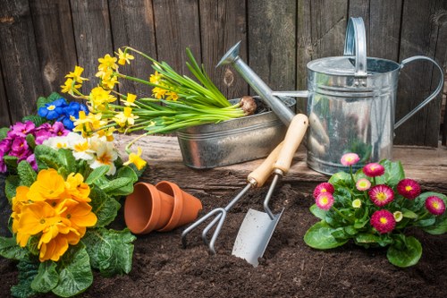 A detailed view of a gardener's tools and flowering plants arranged in a garden bed. In the foreground, vibrant primulas in shades of yellow, pink, and purple are planted in rich, dark soil. To the left, a cluster of yellow primulas with broad green leaves is visible, alongside a pink primula. A small terracotta plant pot is tipped over on the soil surface. In the middle, a gardening fork and hand trowel with wooden handles rest against the soil, positioned next to a metal watering can with a spout, all set against a weathered wooden fence in the background. Behind the tools, there is a rectangular metal container holding a bunch of daffodil leaves with yellow flowers. The scene suggests an outdoor garden area during daytime with natural lighting, showcasing typical elements of garden maintenance and planting activities, relevant to gardening services in Aldgate, London.