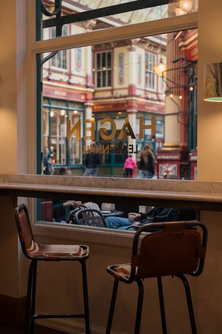 Looking through a large window of a cozy café, the exterior view showcases a charming street scene at Leadenhall Market with ornate, colorful historic buildings and pedestrians walking along the pave
