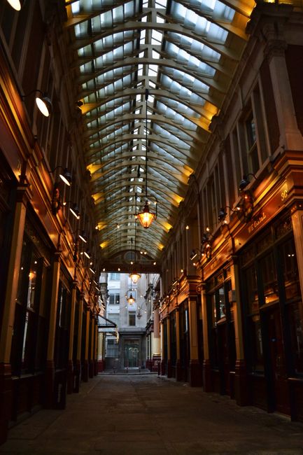 Looking through a large window of a cozy café, the exterior view showcases a charming street scene at Leadenhall Market with ornate, colorful historic buildings and pedestrians walking along the pave